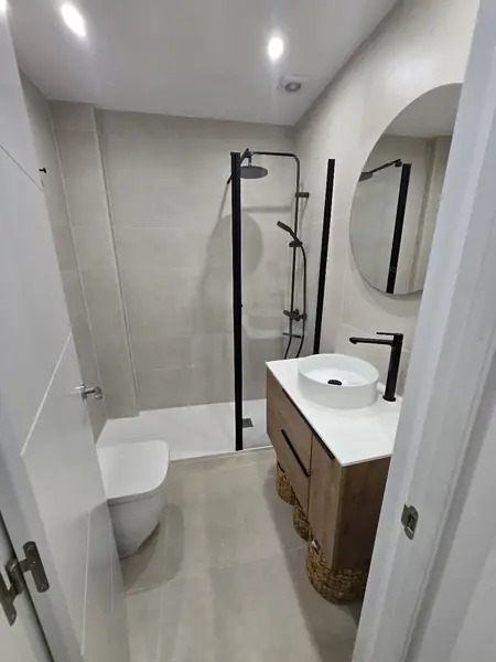 Baño con grifería negra, espejo redondo y mueble suspendido de madera
